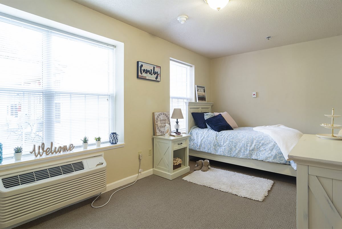 A cozy bedroom with a neatly made bed, bedside table, and natural light from two windows.