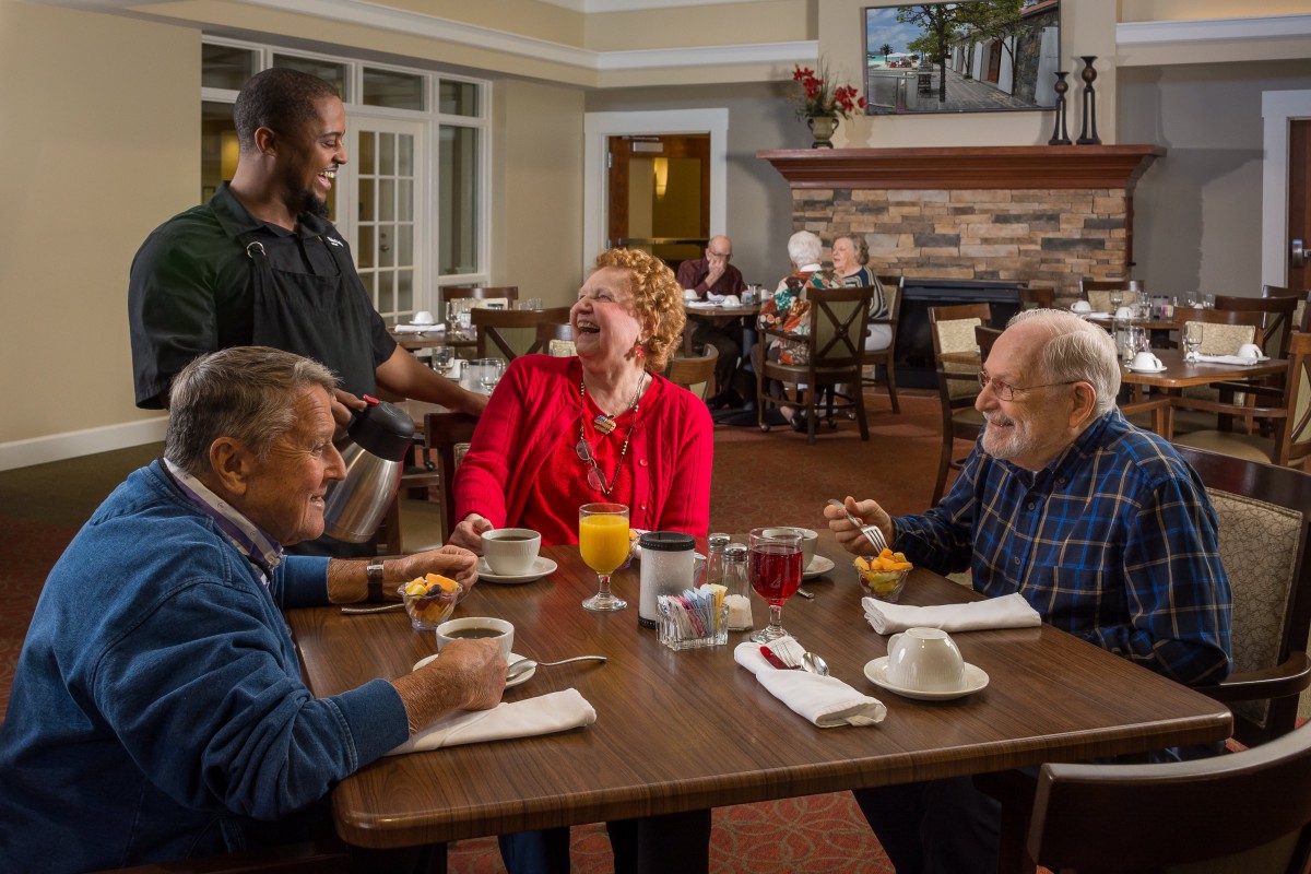 Three seniors enjoy a pleasant breakfast conversation with a waiter in a cozy dining setting.