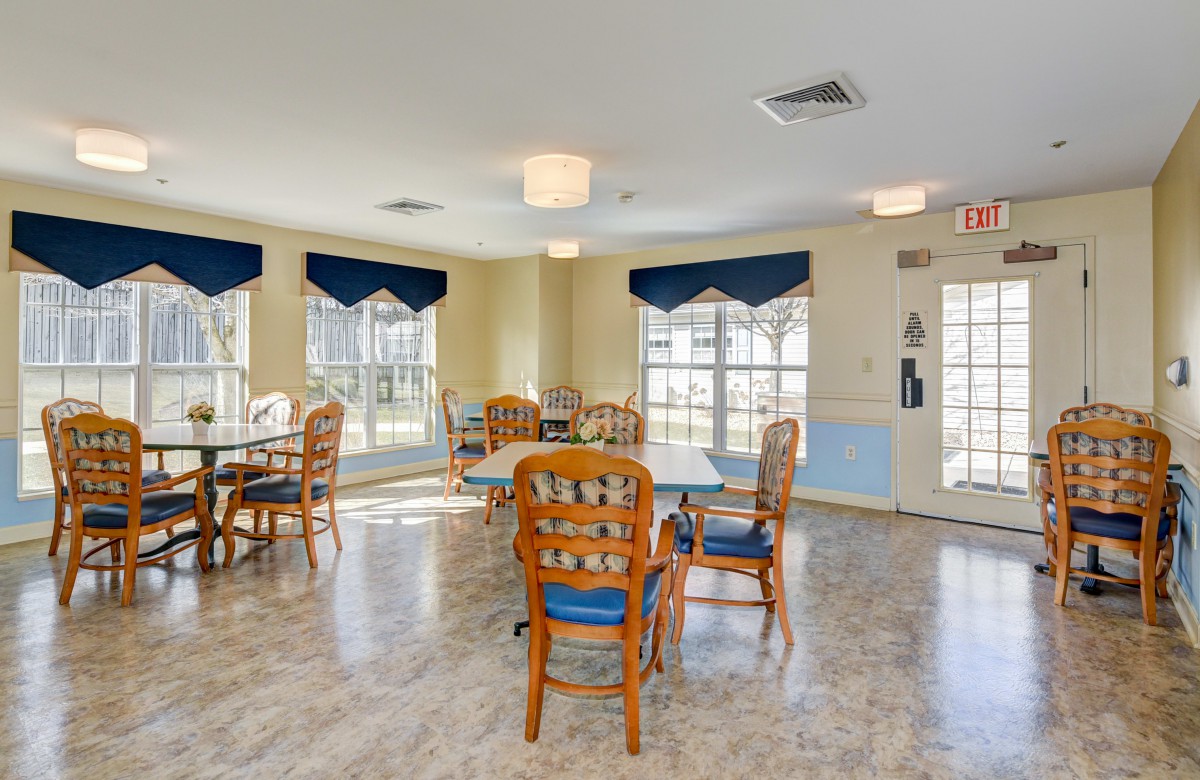 A bright dining room with wooden chairs and tables, featuring large windows with blue curtains.