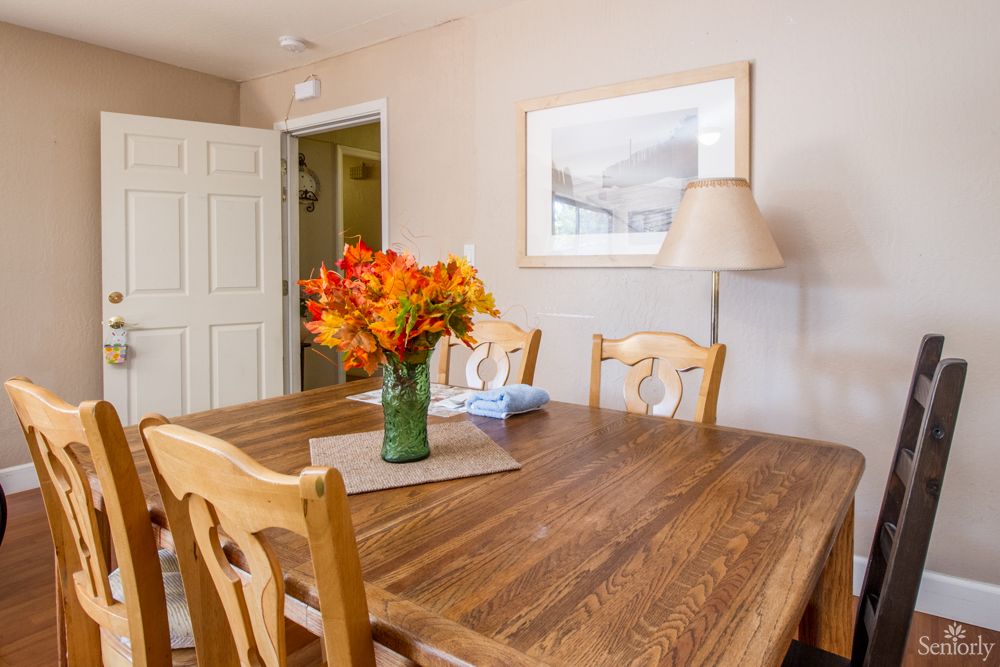 A cozy dining room featuring a wooden table adorned with a vase of vibrant orange flowers, surrounded by matching chairs and complemented by soft lighting and wall art.