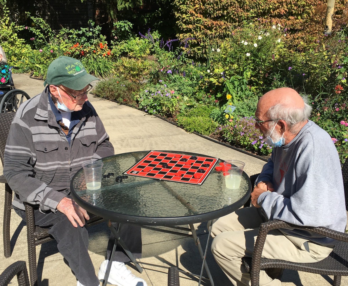 Two elderly men play a game of checkers outdoors in a garden setting on a sunny day.