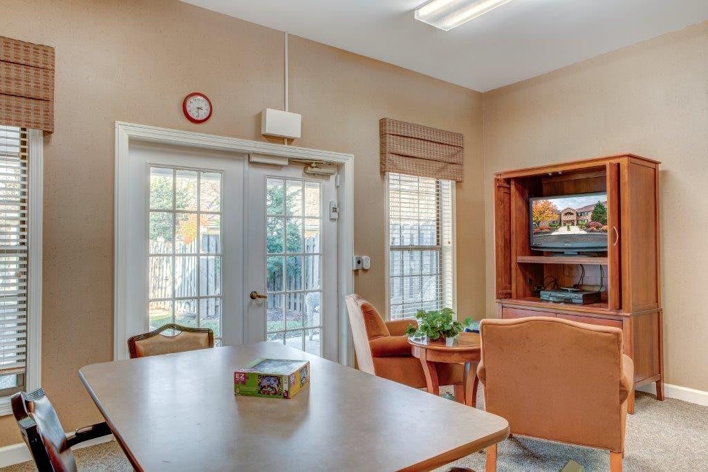 A cozy living room with a table, chairs, and a television near bright windows and a glass door.