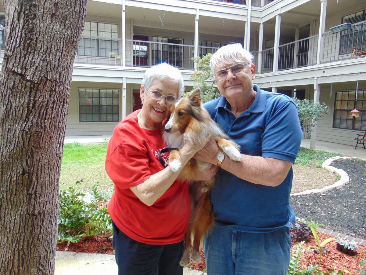 An elderly couple holding a dog and smiling outdoors.