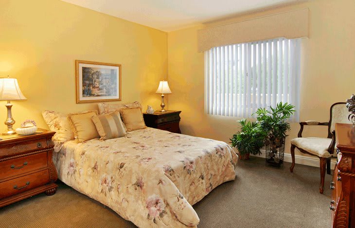 A cozy bedroom with a warm color scheme, featuring a bed with floral bedding, a bedside lamp, and a window with blinds.