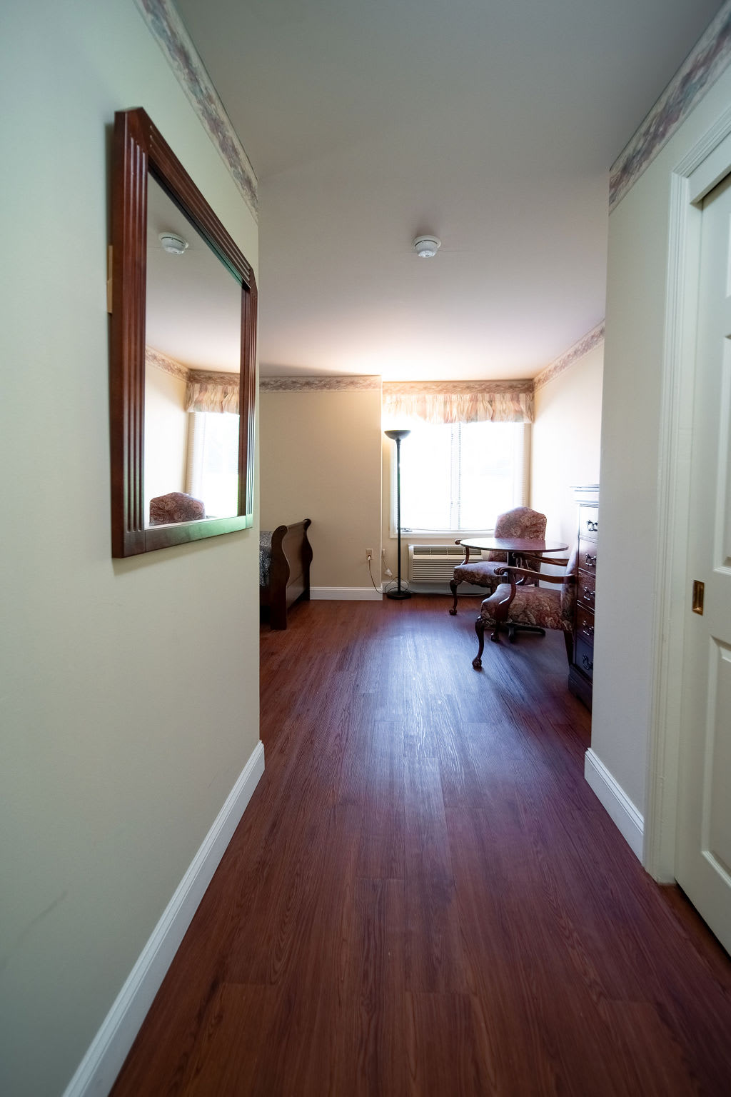 A well-lit hallway featuring a large mirror, wooden flooring, and furniture near the window.