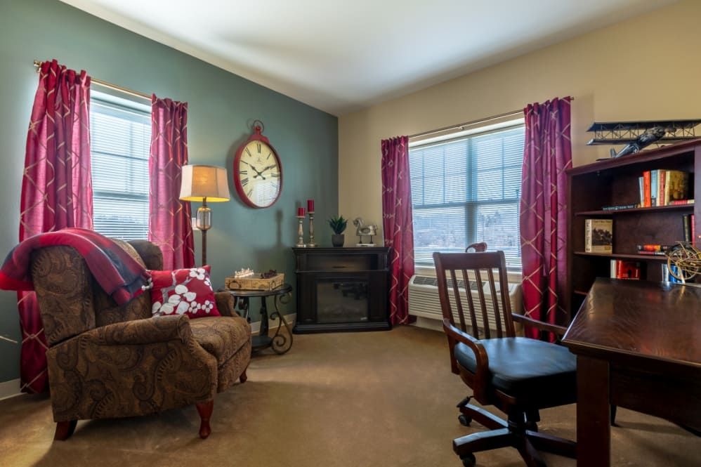 A cozy living room featuring a comfortable armchair, rocking chair, and fireplace, with red curtains and a warm atmosphere.