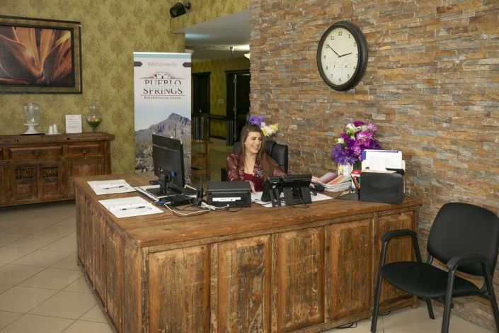 A receptionist sits at a wooden front desk in a hotel lobby, with decor including a clock and flower arrangements.