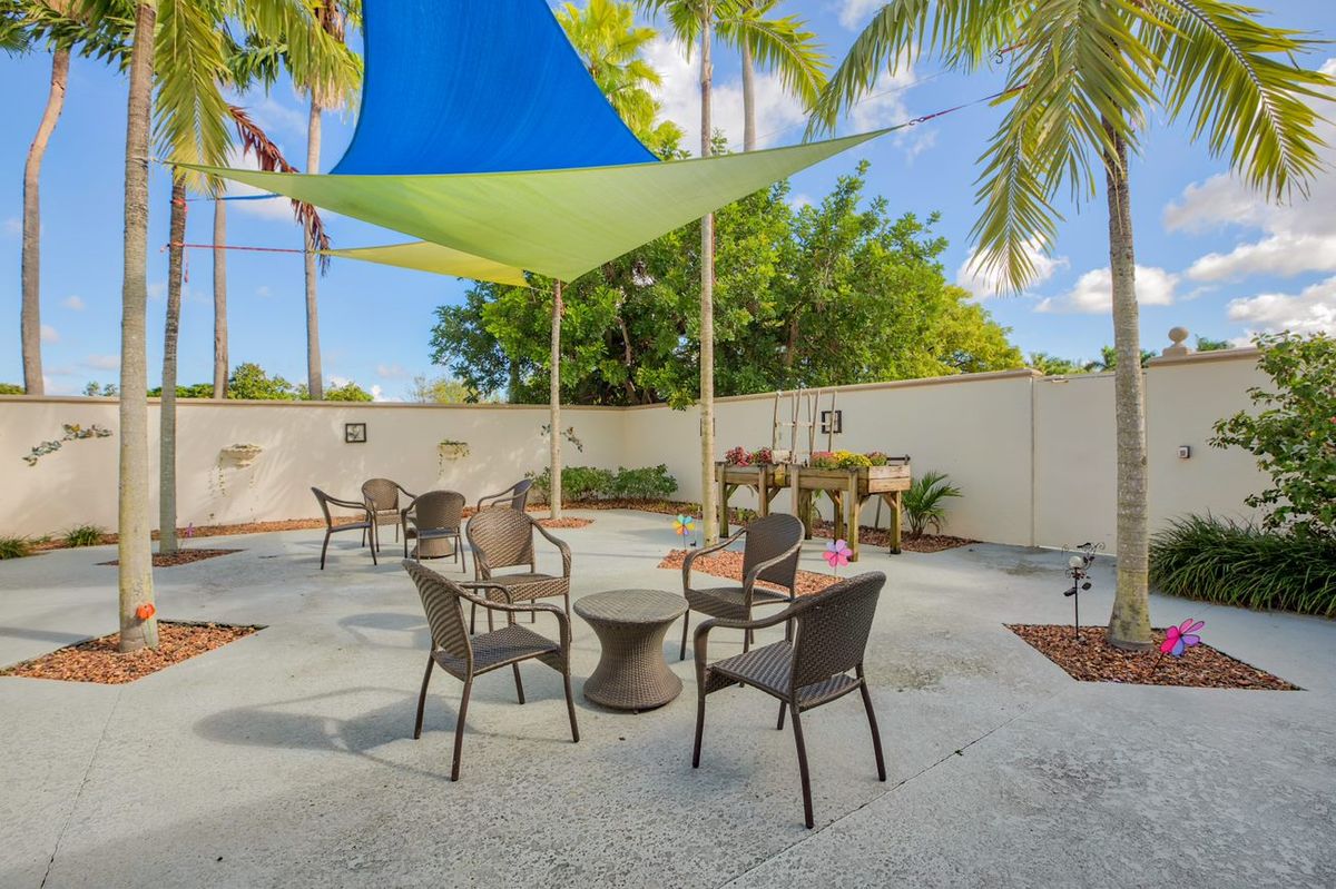 A sunny outdoor patio area with chairs and tables under a colorful shade sail, surrounded by palm trees.