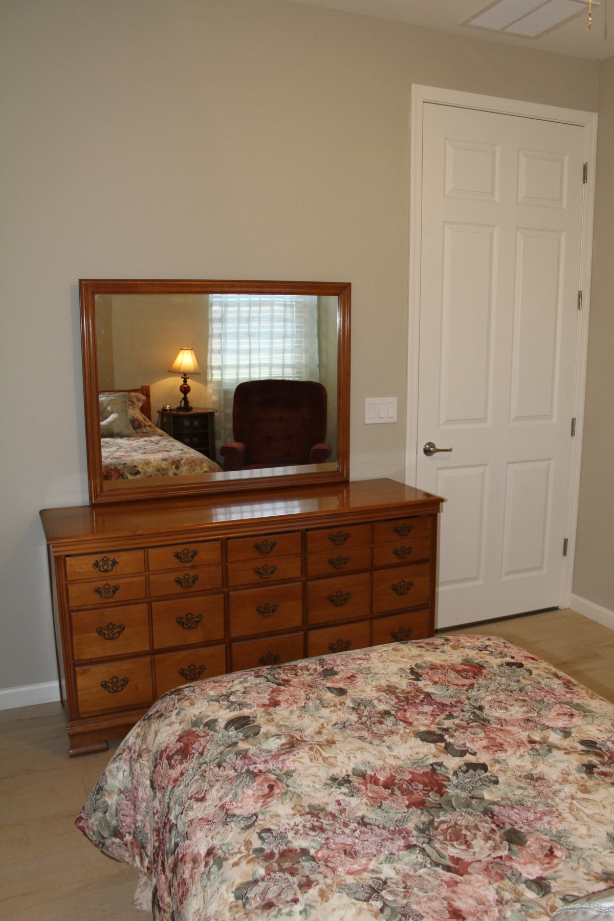 A cozy bedroom with a wooden dresser topped by a large mirror, next to a bed with a floral bedspread.