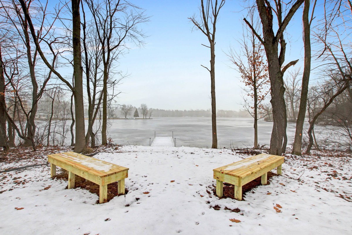 Two benches face a frozen lake surrounded by bare trees on a snowy winter day.