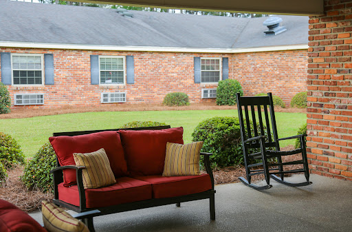 A cozy outdoor seating area featuring a red couch and a rocking chair, with a lush green lawn and a brick house in the background.