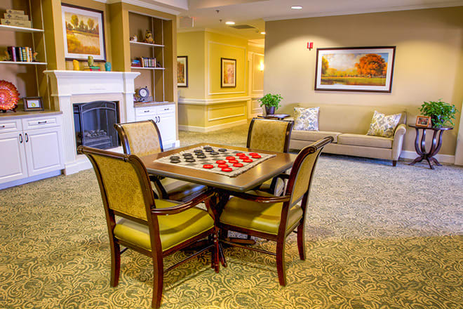 A cozy living room featuring a checkers board set up on a table surrounded by chairs, with a fireplace and artwork in the background.
