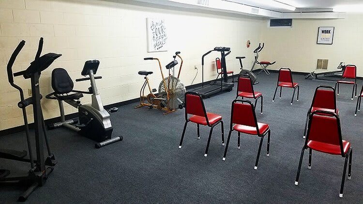 A small gym room with exercise equipment including stationary bikes and a treadmill, alongside a few red chairs.