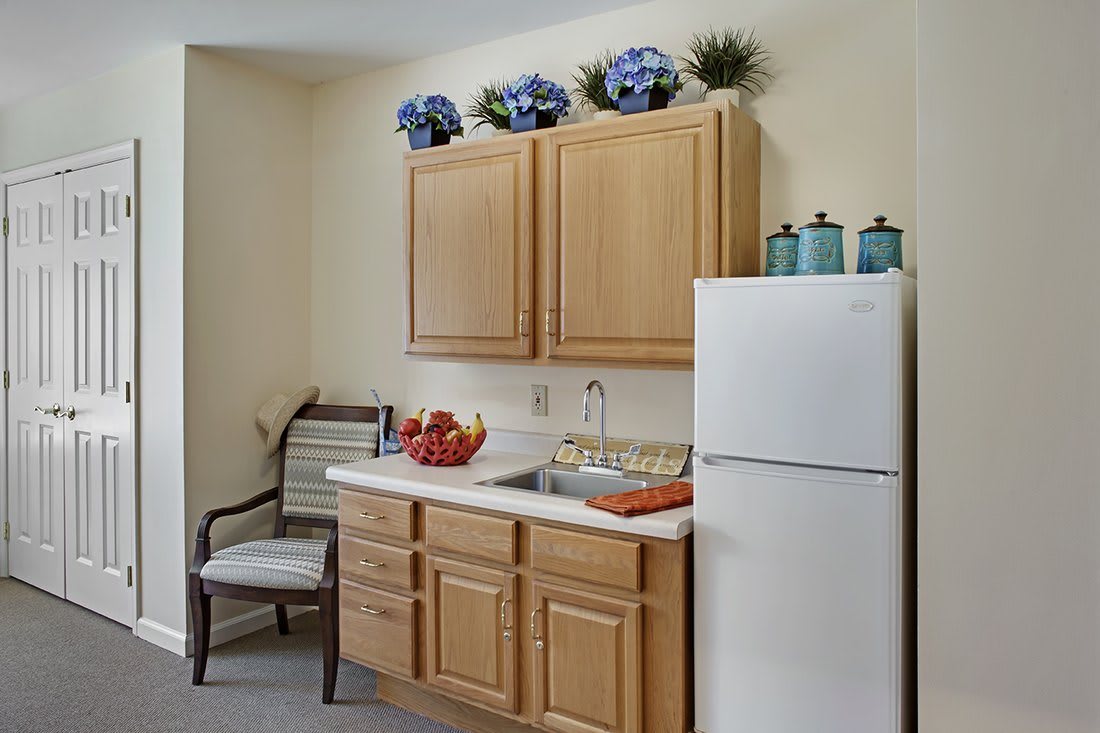 A cozy kitchen corner with wooden cabinets, a sink, and a white refrigerator. Decorative flowers adorn the top of the cabinets.