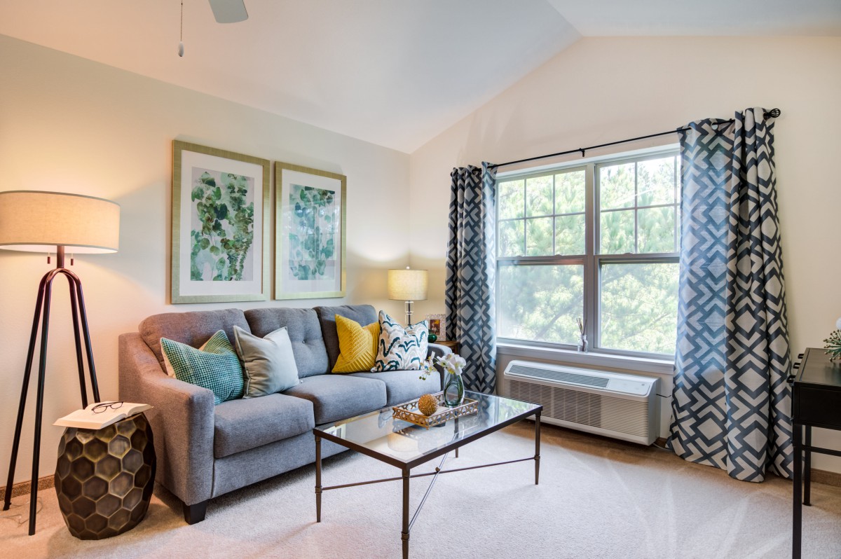 A cozy living room with a comfortable sofa, decorative pillows, and a glass coffee table in front of a large window adorned with patterned curtains.