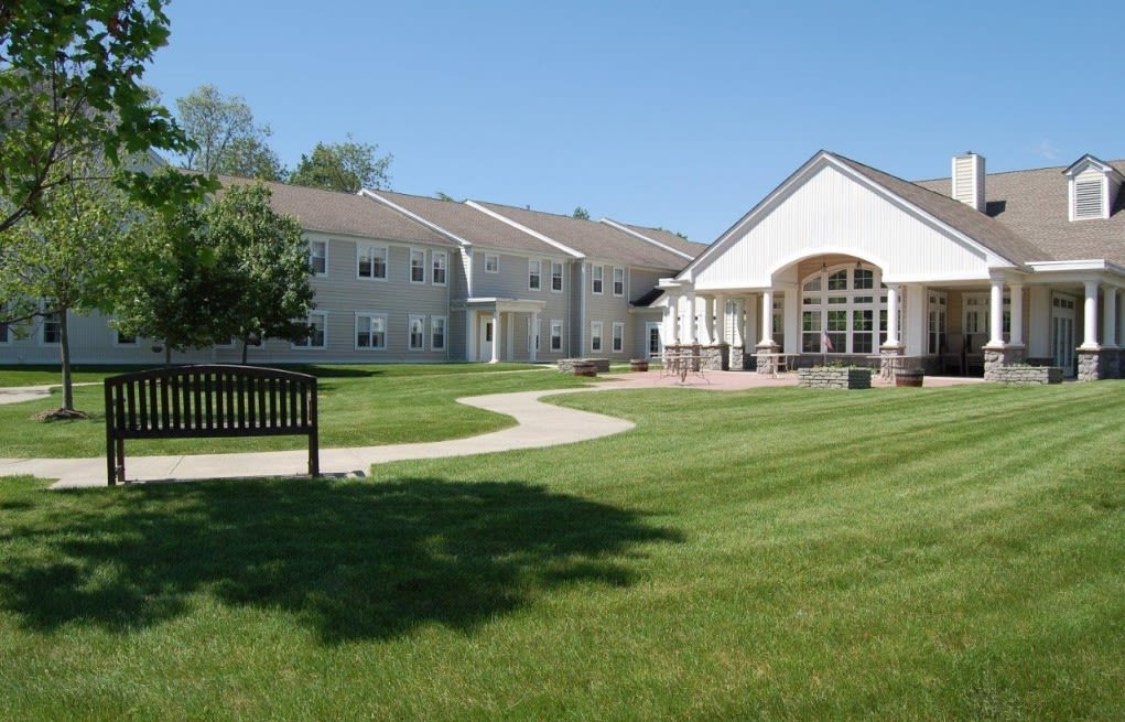 A peaceful residential building surrounded by a green lawn and trees with a bench along a curved pathway.