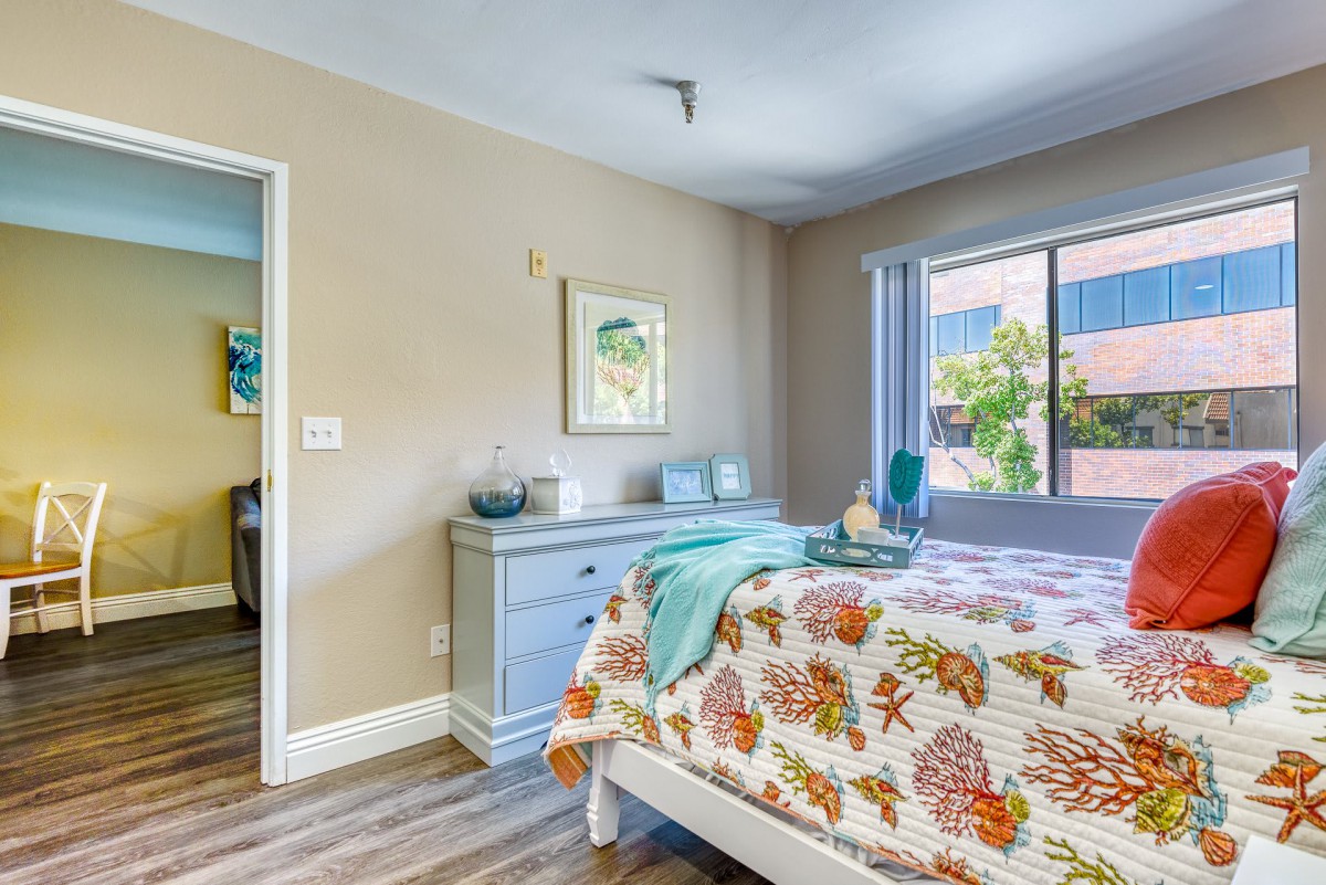 A cozy bedroom with floral bedding, featuring a dresser, artwork on the wall, and a large window offering natural light.