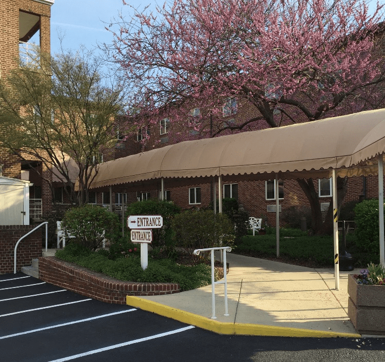 A building entrance with an awning, surrounded by blooming trees and a small parking area.