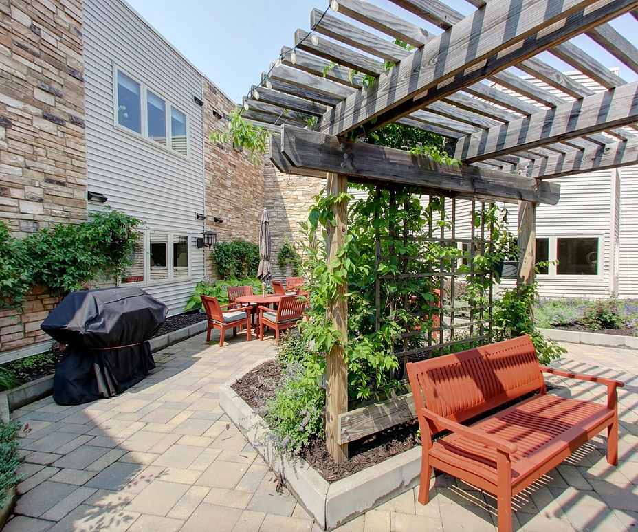A charming outdoor patio with a pergola, featuring wooden benches and chairs surrounded by lush greenery.