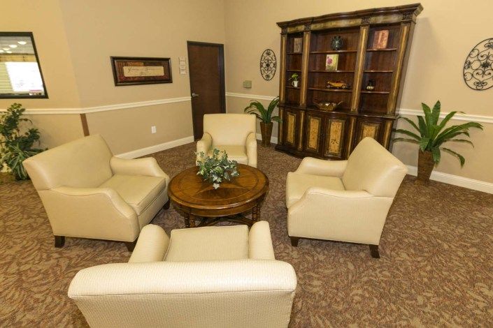 A cozy living room setup featuring four beige armchairs arranged around a round wooden table, with a bookshelf in the background.