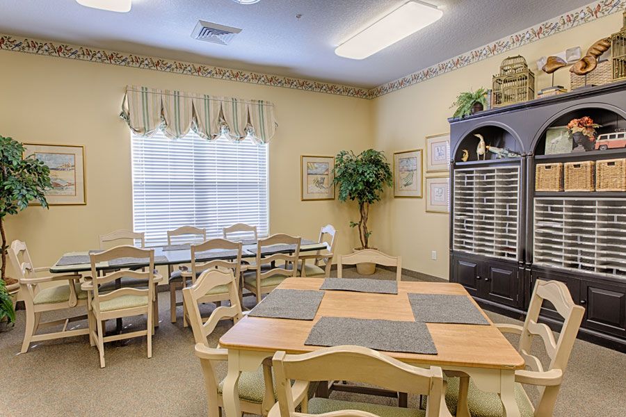 A cozy dining room with several tables and chairs set near a window with curtains, featuring decorative plants and warm lighting.
