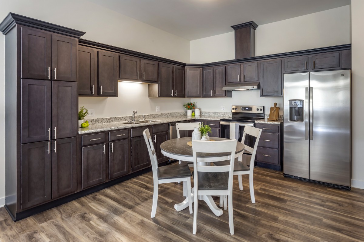A modern kitchen with dark cabinets and stainless steel appliances, featuring a round dining table and chairs on a hardwood floor.