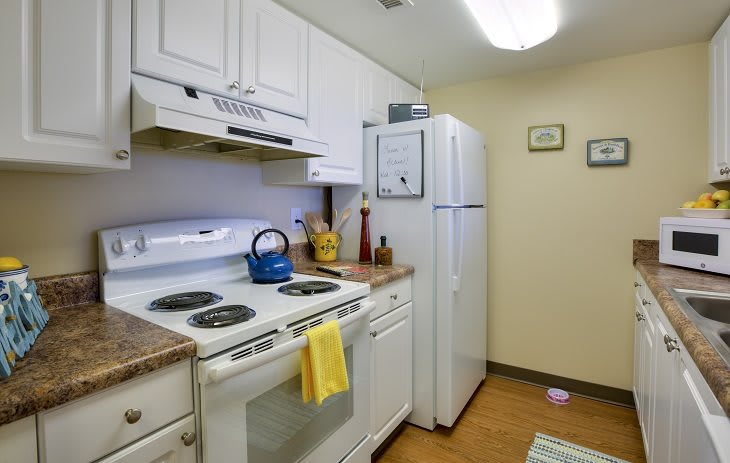 A compact kitchen with white cabinets, featuring a stove, refrigerator, and a blue teapot.