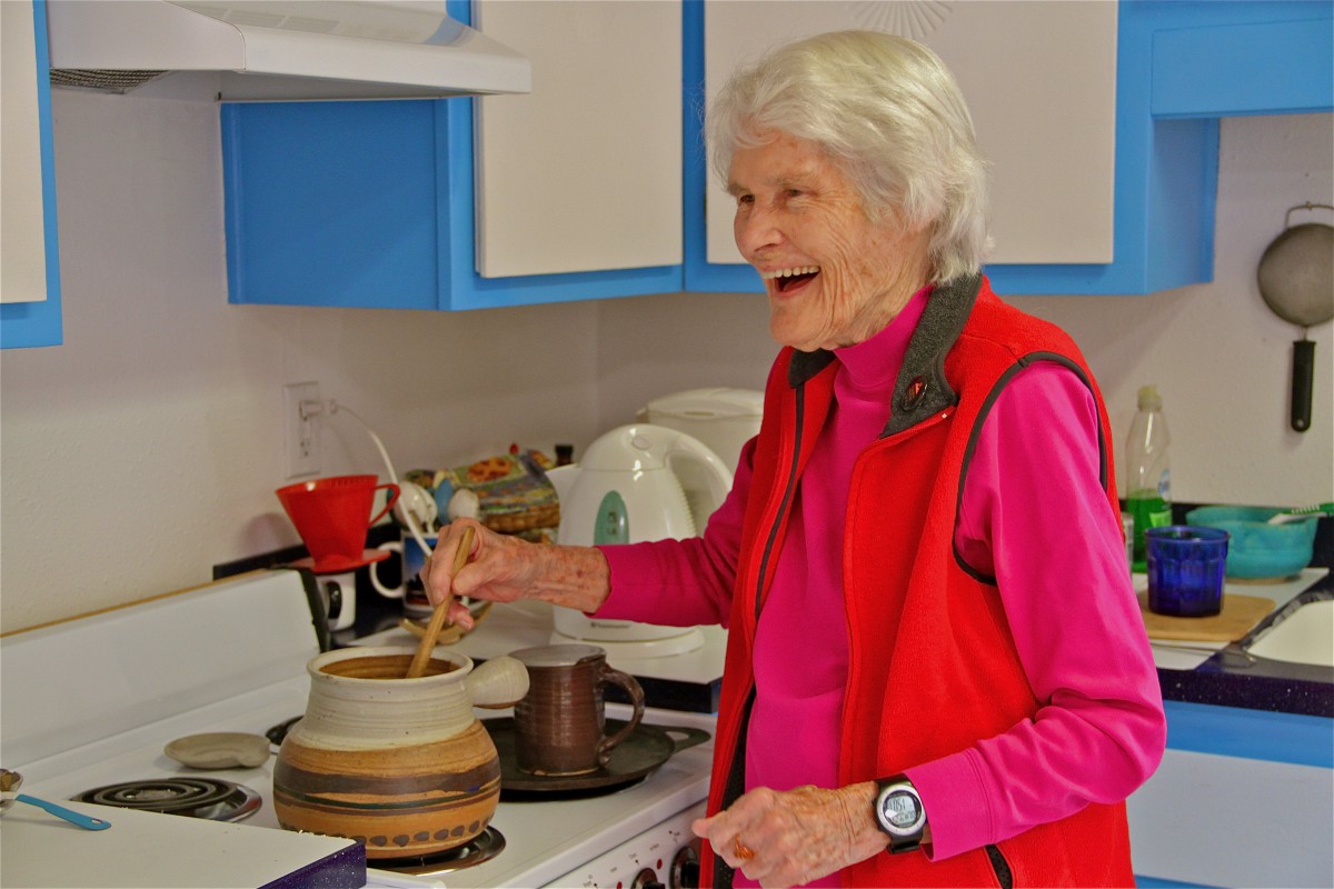An elderly woman smiles while cooking in the kitchen.