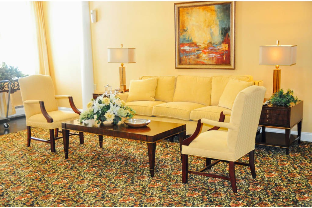 A cozy living room with a yellow sofa, two armchairs, and a wooden coffee table adorned with flowers, complemented by a patterned rug and a lamp next to abstract wall art.