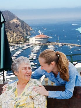 An elderly woman and a young woman smiling together with a marina and mountains in the background.