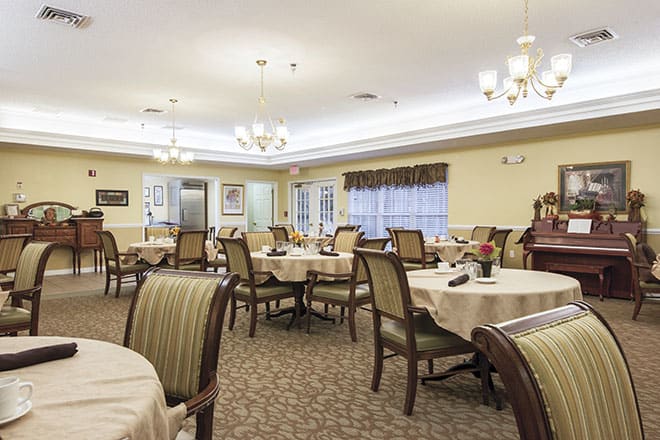 A spacious dining room with elegantly set tables and upholstered chairs, illuminated by chandeliers.