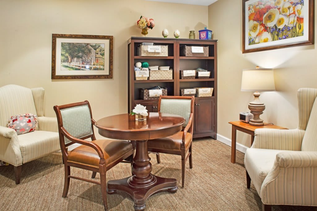A cozy office interior featuring a round wooden table, two chairs, a bookshelf with neatly organized items, and tasteful artwork on the walls.