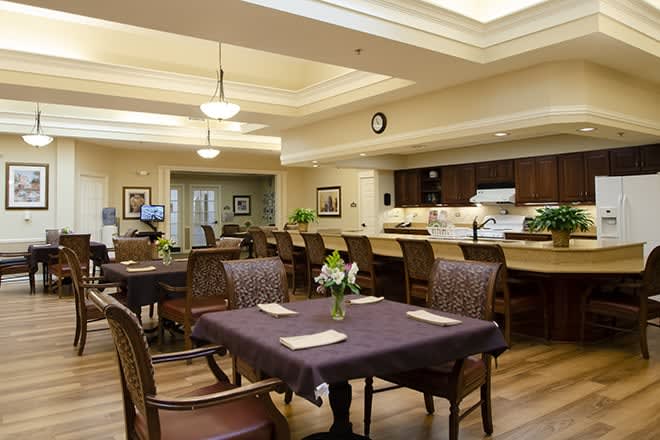A spacious dining area with a kitchen in the background, featuring tables and chairs under warm ceiling lighting.