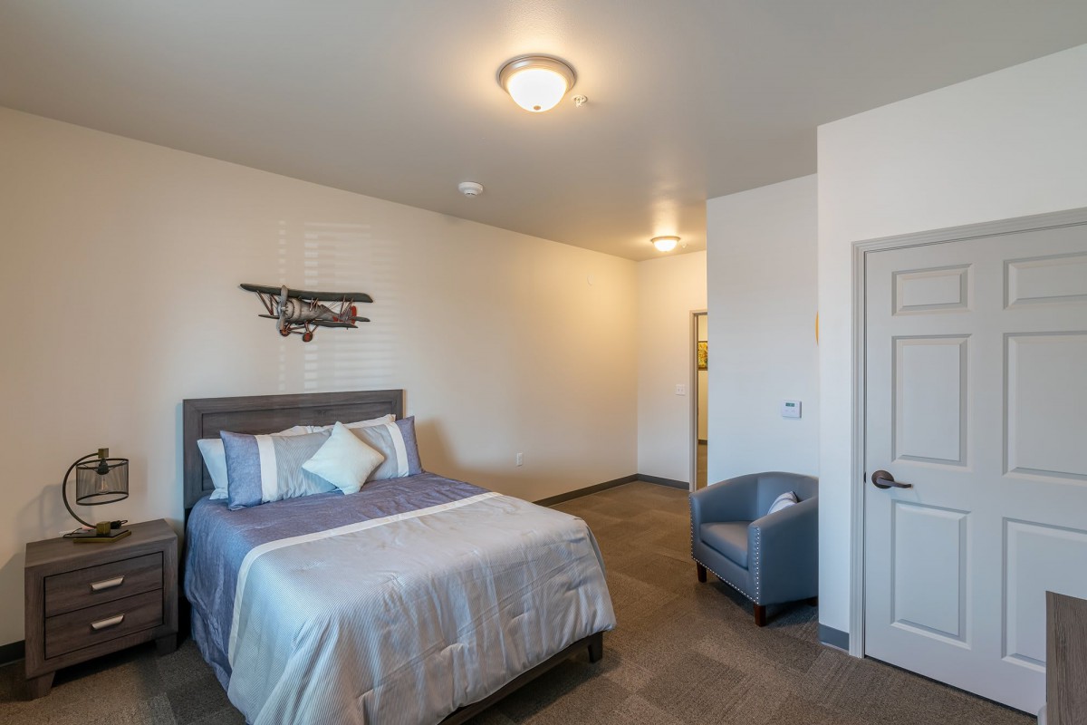 A modern bedroom with a neatly made bed, an armchair, and wall art in neutral tones.