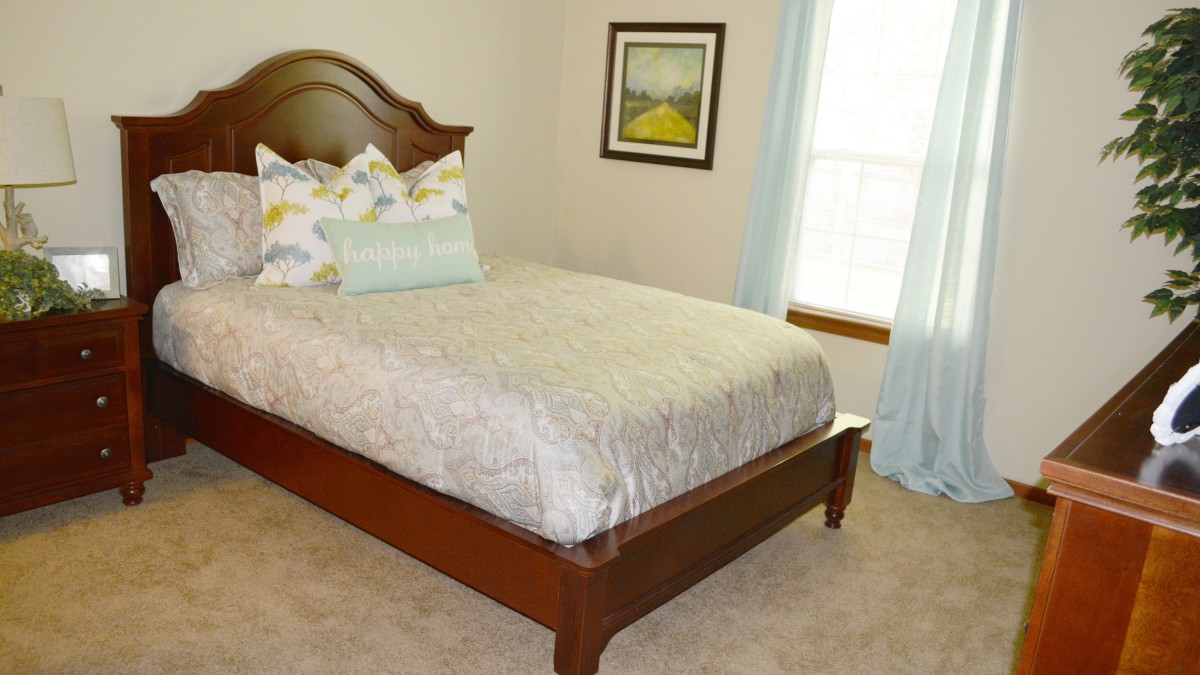 A cozy bedroom featuring a neatly made bed with decorative pillows, soft carpeting, and a framed artwork on the wall.