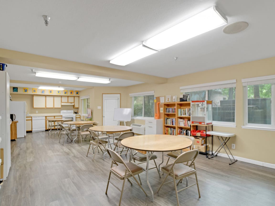 A spacious classroom with round tables, folding chairs, bookshelves, and bright overhead lighting.