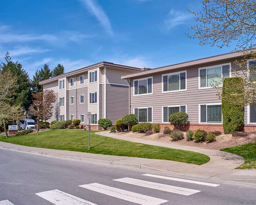 A well-maintained apartment building with neatly landscaped grounds and a crosswalk in the foreground under a clear blue sky.