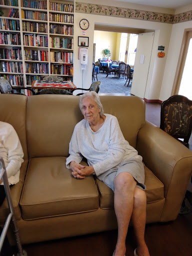 An elderly woman relaxing on a sofa in a communal space with bookshelves in the background.
