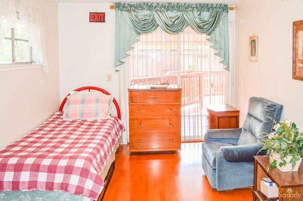A cozy bedroom with a single plaid-bedded frame, dresser, and comfortable armchair next to a window with curtains.