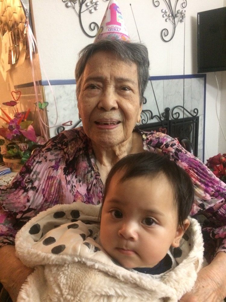 An elderly woman holding a baby, both smiling indoors.