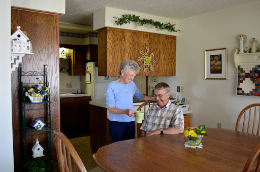 An elderly woman assists a man seated at the dining table in a cozy kitchen setting.