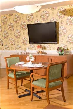 A cozy dining area featuring a round table with chairs, a cabinet, and a wall-mounted television against floral wallpaper.