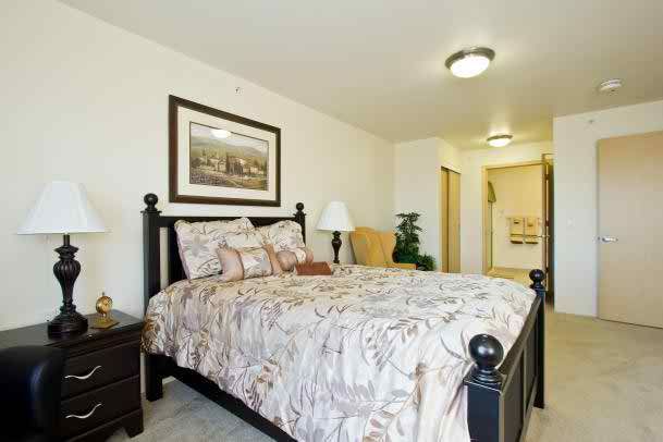 A cozy bedroom featuring a neatly made bed with a patterned comforter, a nightstand with a lamp, and artwork adorning the wall.