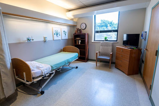 A clean and well-organized hospital room featuring a bed, chair, and natural light from the window.