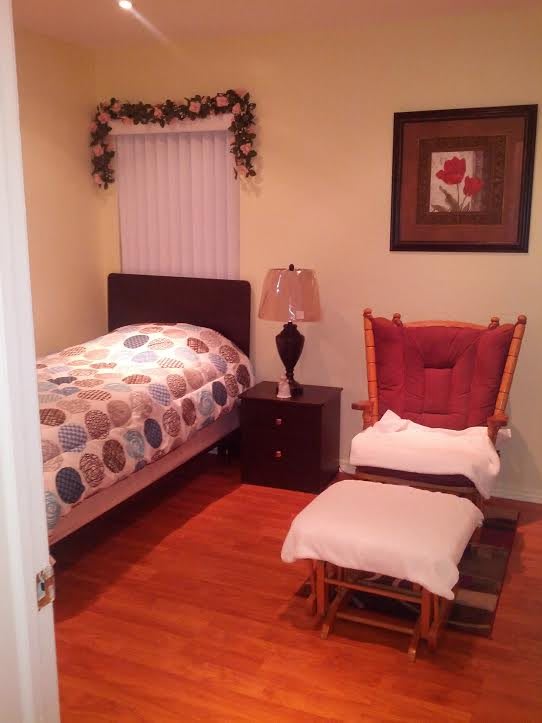 A cozy bedroom with a bed, nightstand, lamp, and a cushioned chair with a footrest.