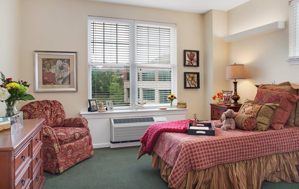 A cozy bedroom with a large window, an armchair, and a neatly made bed with a red bedspread.