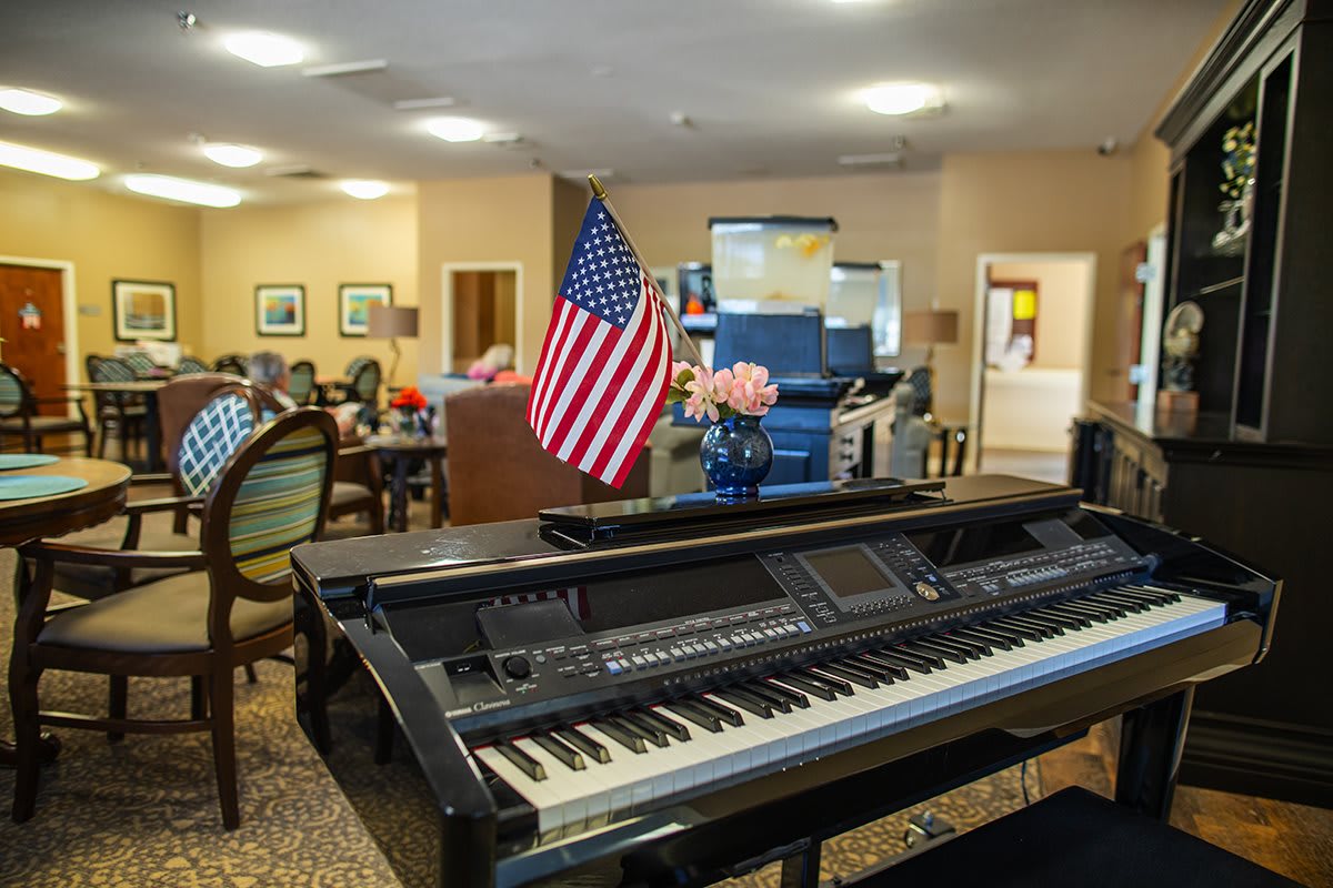 A piano adorned with an American flag in a community center with people sitting and flowers on display.