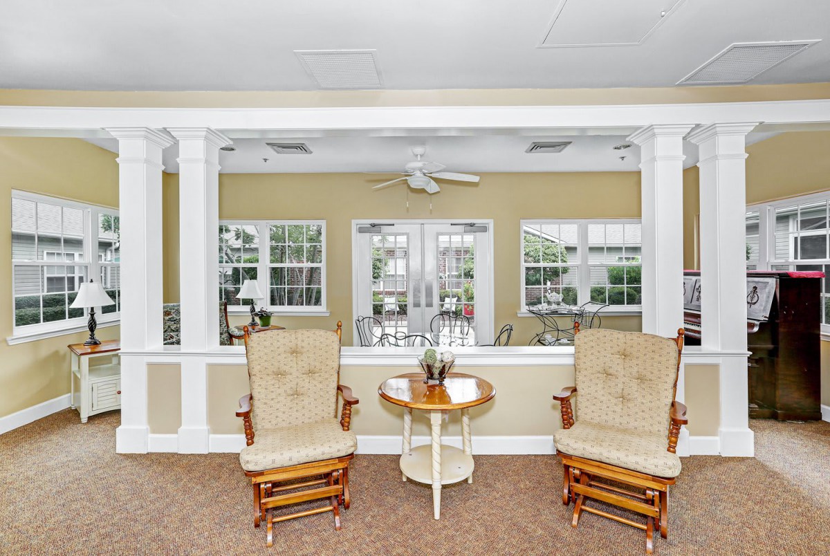A cozy sitting area with two cushioned chairs and a small round table, set against large windows and a ceiling fan.