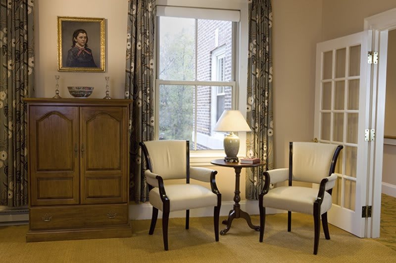 A cozy living room corner featuring elegant chairs, a lamp on a side table, and a large window with curtains.