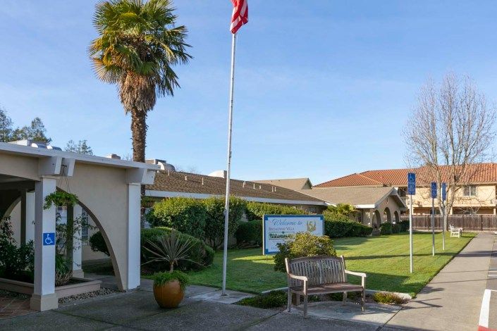 A building with a flagpole and garden, featuring a bench and palm tree under a clear blue sky.
